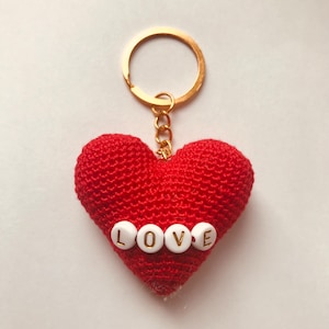 Puede incluir: Llavero de coraz&oacute;n rojo tejido a crochet con una cadena dorada y un anillo para llaves. El coraz&oacute;n tiene cuentas blancas con letras doradas que deletrean "LOVE".