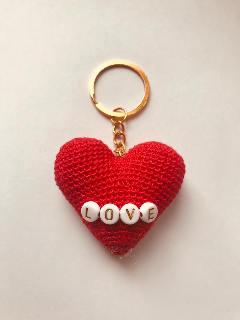 Puede incluir: Llavero de coraz&oacute;n rojo tejido a crochet con una cadena dorada y un anillo para llaves. El coraz&oacute;n tiene cuentas blancas con letras doradas que deletrean "LOVE".