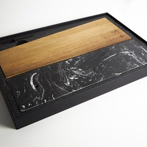 Puede incluir: Una tabla de servir rectangular con un dise&ntilde;o dividido, compuesta por una mitad de madera marr&oacute;n clara y otra mitad de piedra negra con vetas blancas similares al m&aacute;rmol, presentada dentro de una caja de almacenamiento negra con espuma.