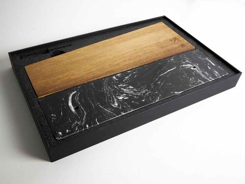 Puede incluir: Una tabla de servir rectangular con un dise&ntilde;o dividido, compuesta por una mitad de madera marr&oacute;n clara y otra mitad de piedra negra con vetas blancas similares al m&aacute;rmol, presentada dentro de una caja de almacenamiento negra con espuma.