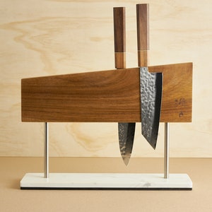 May include: A magnetic knife block made of dark wood with a horizontal grain, supported by two silver metal posts on a rectangular white marble base. Two kitchen knives with hammered metal blades and dark wooden handles are attached to the block.