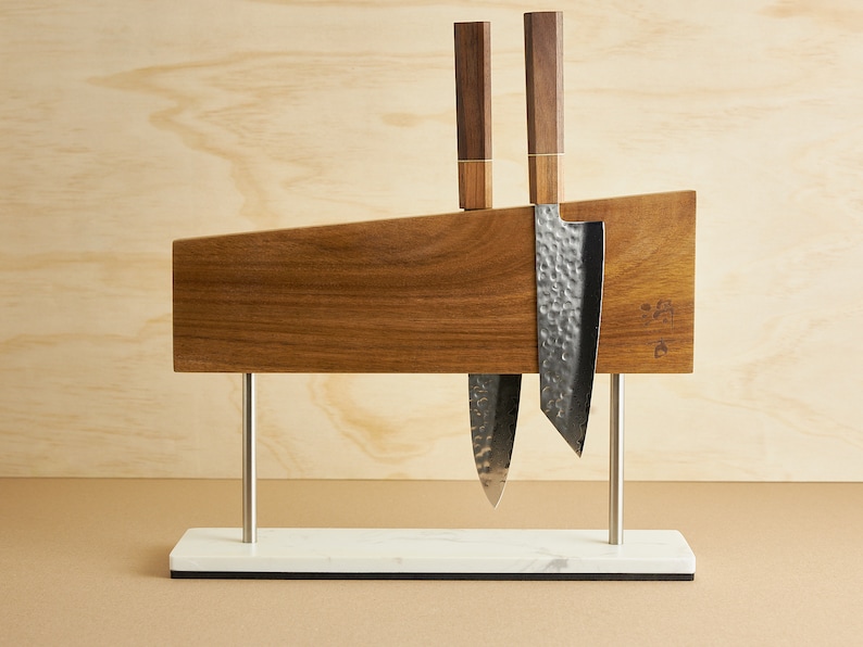 May include: A magnetic knife block made of dark wood with a horizontal grain, supported by two silver metal posts on a rectangular white marble base. Two kitchen knives with hammered metal blades and dark wooden handles are attached to the block.