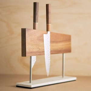 May include: A magnetic knife block featuring a rectangular wooden board supported by two silver metal rods on a white marble base. Two kitchen knives with dark wooden handles and hammered metal blades are attached to the front of the wooden board.