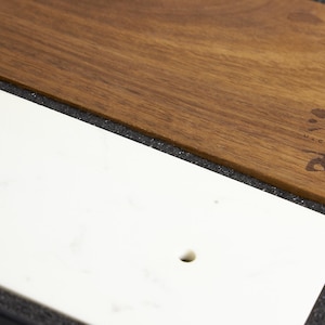 Puede incluir: Una tabla de servir rectangular que presenta una secci&oacute;n de m&aacute;rmol blanco liso con un peque&ntilde;o orificio circular y una secci&oacute;n de madera marr&oacute;n c&aacute;lida con veta oscura. La parte de madera muestra un peque&ntilde;o logotipo oscuro estilizado con el texto u-co.