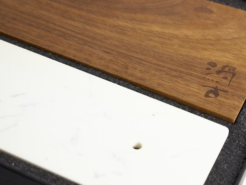 Puede incluir: Una tabla de servir rectangular que presenta una secci&oacute;n de m&aacute;rmol blanco liso con un peque&ntilde;o orificio circular y una secci&oacute;n de madera marr&oacute;n c&aacute;lida con veta oscura. La parte de madera muestra un peque&ntilde;o logotipo oscuro estilizado con el texto u-co.