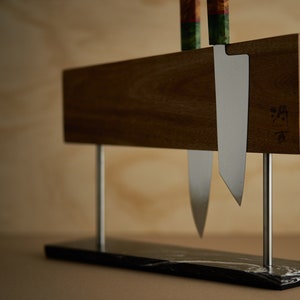 May include: A magnetic knife holder featuring a horizontal wooden block supported by two silver metal rods on a black marble base. Two kitchen knives with colourful patterned handles are attached to the wood, which displays a small engraved logo.