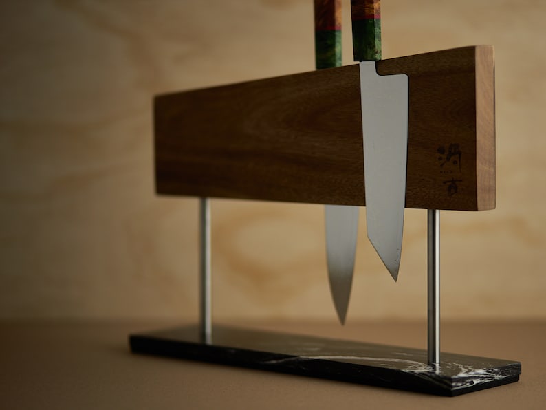 May include: A magnetic knife holder featuring a horizontal wooden block supported by two silver metal rods on a black marble base. Two kitchen knives with colourful patterned handles are attached to the wood, which displays a small engraved logo.