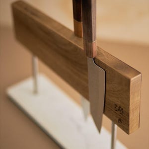 May include: A magnetic knife block featuring a rectangular wooden board supported by two silver metal rods on a white marble base. Two kitchen knives with dark wooden handles are attached to the board, which displays a small dark logo.