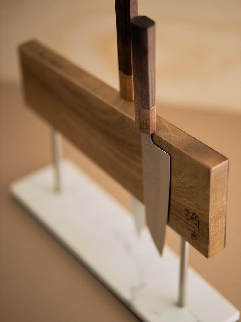 May include: A magnetic knife block featuring a rectangular wooden board supported by two silver metal rods on a white marble base. Two kitchen knives with dark wooden handles are attached to the board, which displays a small dark logo.