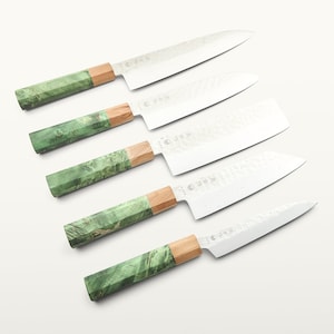 Op de afbeelding: Een set van vijf keukenmessen met zilverkleurige metalen bladen met een gehamerde textuur en Japanse karakters. Elk mes heeft een handvat dat bestaat uit een licht houten gedeelte en een gemarmerd groen harsgedeelte met een achthoekige vorm.