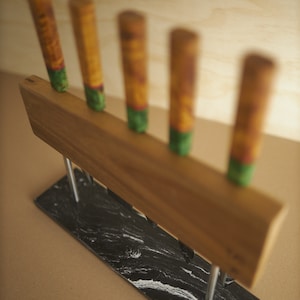 Puede incluir: Un bloque para cuchillos con un soporte rectangular de madera y cinco mangos verticales, cada uno con un patr&oacute;n amarillo y verde. El soporte de madera se apoya en dos varillas met&aacute;licas plateadas fijadas a una base rectangular de m&aacute;rmol blanco y negro.