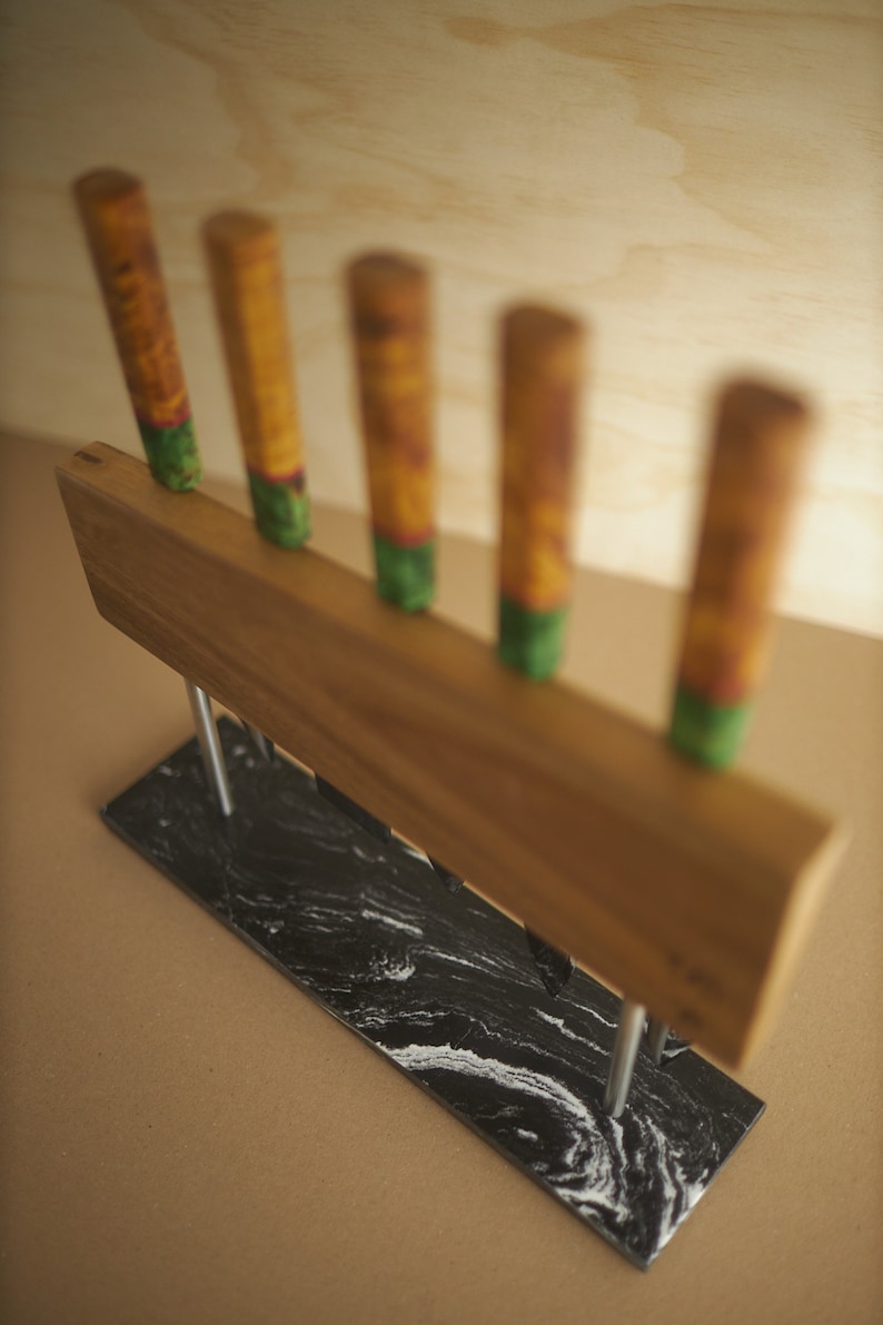 Puede incluir: Un bloque para cuchillos con un soporte rectangular de madera y cinco mangos verticales, cada uno con un patr&oacute;n amarillo y verde. El soporte de madera se apoya en dos varillas met&aacute;licas plateadas fijadas a una base rectangular de m&aacute;rmol blanco y negro.