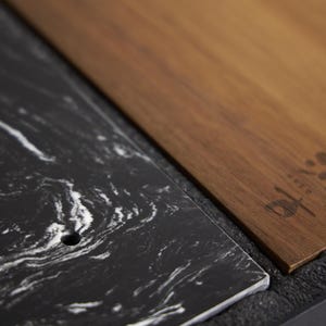 May include: A rectangular black marble slab with white veining and a small circular hole, positioned next to a rectangular brown wooden board featuring a dark engraved logo with the text UECO. The two pieces are set against a dark textured base.