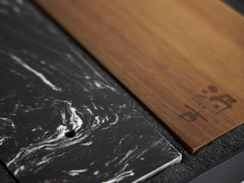 May include: A rectangular black marble slab with white veining and a small circular hole, positioned next to a rectangular brown wooden board featuring a dark engraved logo with the text UECO. The two pieces are set against a dark textured base.