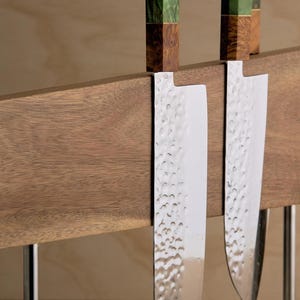 May include: Two kitchen knives with hammered steel blades and handles featuring green and brown wood grain patterns, held by a horizontal wooden magnetic block stand with a small Japanese character inscription on the right side.