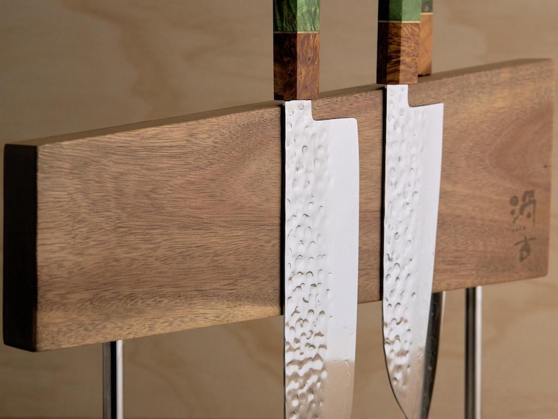 May include: Two kitchen knives with hammered steel blades and handles featuring green and brown wood grain patterns, held by a horizontal wooden magnetic block stand with a small Japanese character inscription on the right side.