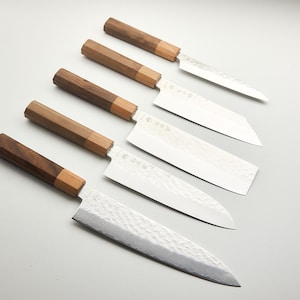 Può includere: Un assortimento di sei coltelli in stile giapponese con manici in legno. I manici sono una combinazione di legno marrone scuro e marrone chiaro. Le lame sono argentate e hanno una finitura strutturata. I coltelli sono disposti in diagonale.