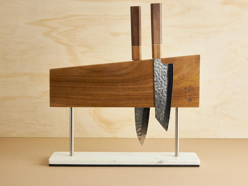 Puede incluir: Un bloque magn&eacute;tico de madera con veta rectangular, sostenido por dos varillas de metal plateado sobre una base de m&aacute;rmol blanco. Dos cuchillos de cocina con mangos de madera oscura y hojas de metal martillado est&aacute;n sujetos a la superficie.