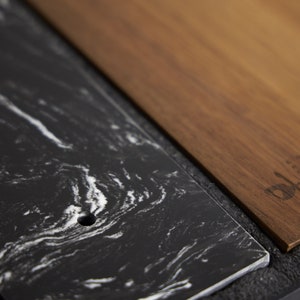 Puede incluir: Una losa rectangular de m&aacute;rmol negro con vetas blancas y un orificio circular, colocada junto a una tabla rectangular de madera marr&oacute;n con un logotipo oscuro grabado que dice Meco. Ambos elementos est&aacute;n en un marco negro texturizado.