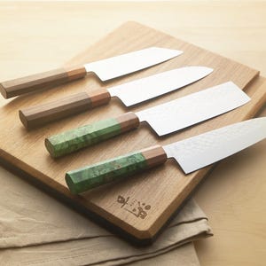 Puede incluir: Cuatro cuchillos de cocina con mangos de madera y verdes sobre una tabla de cortar de madera. Las hojas son plateadas y los mangos son marrones y verdes. Un pa&ntilde;o beige est&aacute; en primer plano.