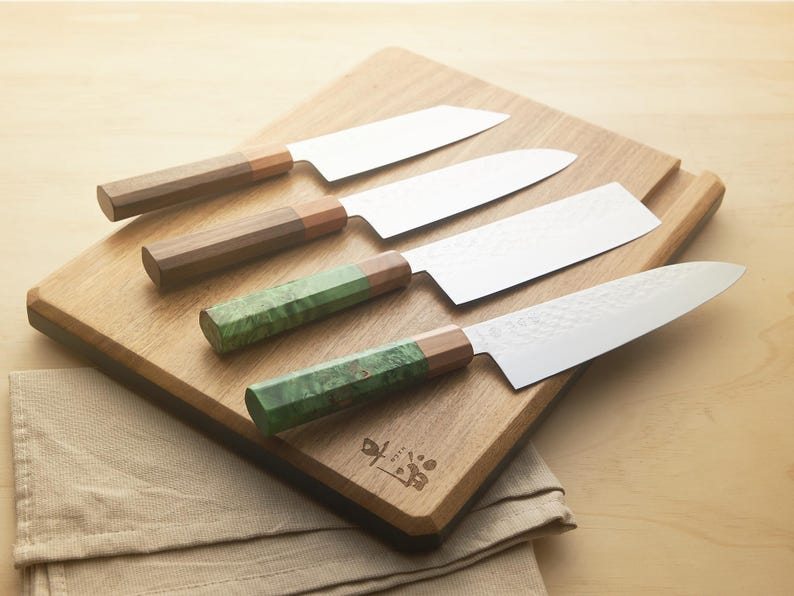 Puede incluir: Cuatro cuchillos de cocina con mangos de madera y verdes sobre una tabla de cortar de madera. Las hojas son plateadas y los mangos son marrones y verdes. Un pa&ntilde;o beige est&aacute; en primer plano.