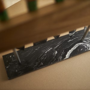 Puede incluir: Una base rectangular de m&aacute;rmol blanco y negro con una superficie lisa y pulida que presenta patrones en espiral. Dos varillas met&aacute;licas verticales se extienden hacia arriba desde la piedra, sosteniendo un bloque de madera horizontal.
