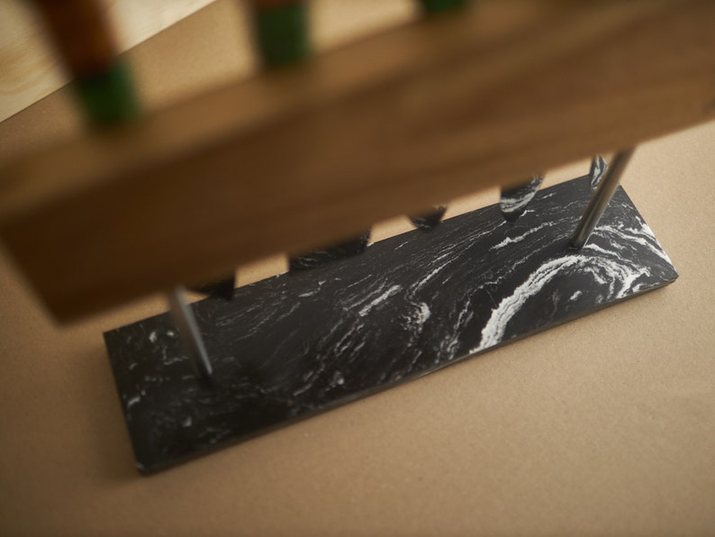 Puede incluir: Una base rectangular de m&aacute;rmol blanco y negro con una superficie lisa y pulida que presenta patrones en espiral. Dos varillas met&aacute;licas verticales se extienden hacia arriba desde la piedra, sosteniendo un bloque de madera horizontal.