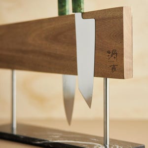 May include: A magnetic knife holder featuring a horizontal walnut wood block with a stamped logo, supported by two silver metal rods attached to a rectangular black marble base with white veining, holding two knives with patterned handles.