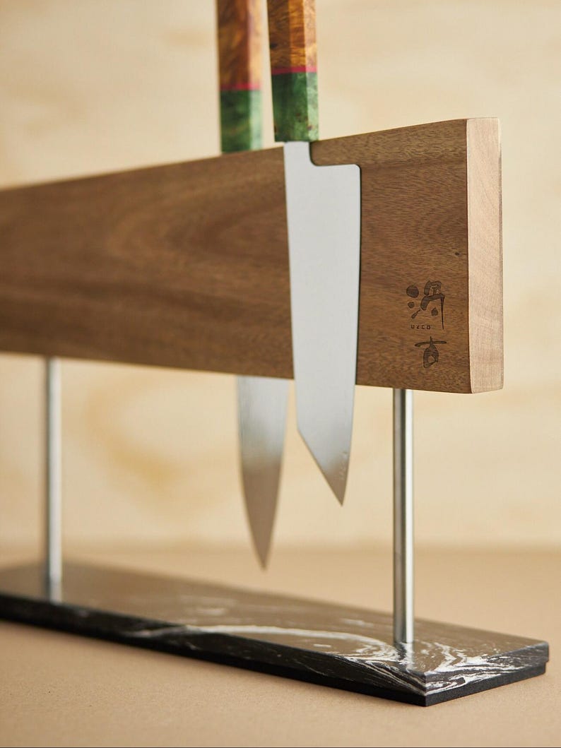 May include: A magnetic knife holder featuring a horizontal walnut wood block with a stamped logo, supported by two silver metal rods attached to a rectangular black marble base with white veining, holding two knives with patterned handles.