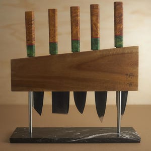 May include: A magnetic knife block featuring a horizontal wooden board with five knives suspended by their blades. The handles are made of light brown wood with green accents. The block is supported by two silver metal rods on a black marble base.
