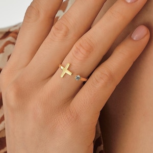 May include: A gold-colored ring featuring a cross design and a small, round, gray stone. The ring is open at the band, with the cross on one side and the stone on the other. The ring is worn on a finger.