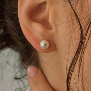 May include: A close-up of a pearl stud earring on an ear. The earring is a small, round, white pearl. The person's skin is fair, and their hair is dark brown and wavy.