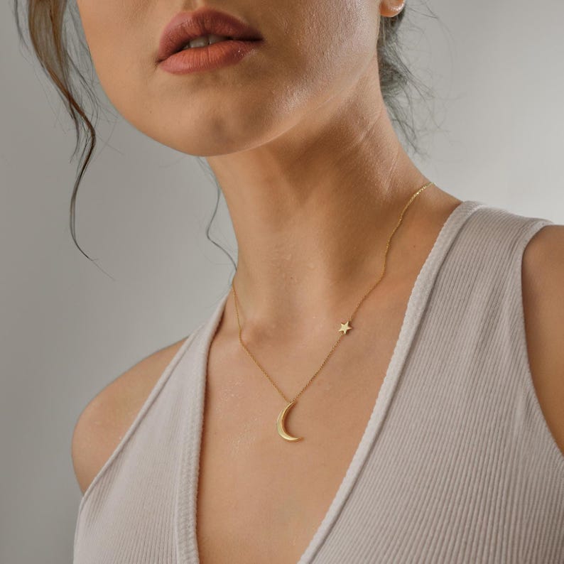 Pu&ograve; includere: Una collana d'oro con un ciondolo a forma di luna crescente e un piccolo ciondolo a forma di stella. La collana &egrave; indossata da una persona con i capelli scuri e una canotta bianca.