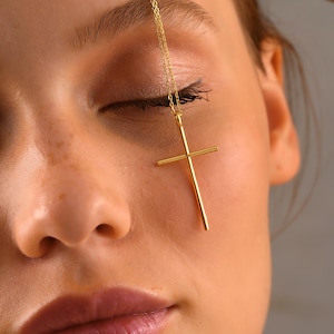 May include: A gold-colored cross pendant necklace. The cross is suspended from a delicate chain. The necklace is shown against a person's face, with the cross resting near the eye.