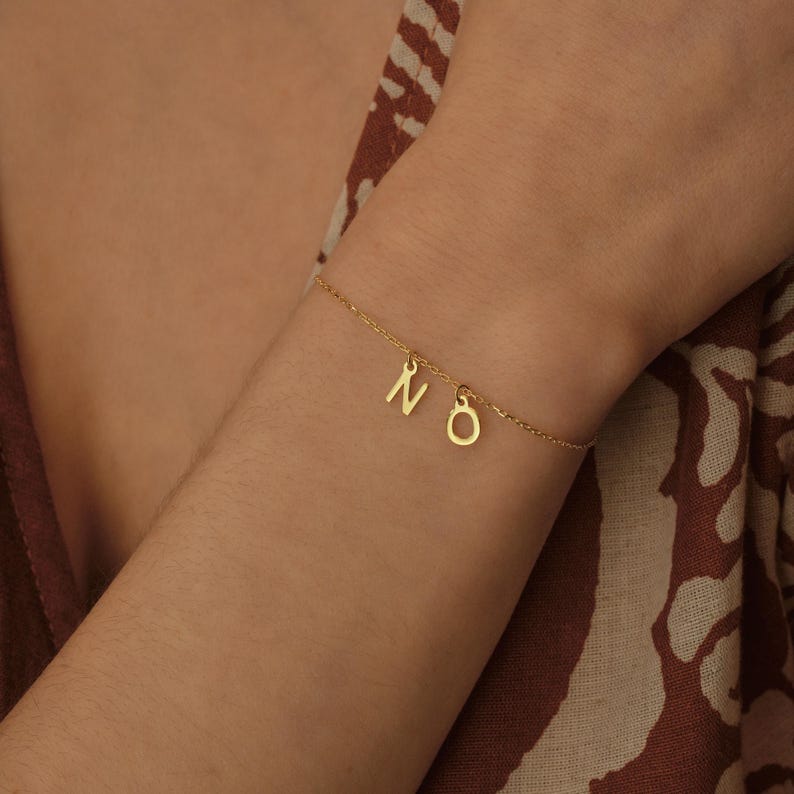 K&ouml;nnte beinhalten: Ein goldenes Kettenarmband mit zwei Anh&auml;ngern, einem Buchstaben N und einem Buchstaben O. Das Armband befindet sich an einem Handgelenk.