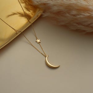 Pu&ograve; includere: Una collana d'oro con un ciondolo a forma di luna crescente e un piccolo ciondolo a forma di stella. La collana &egrave; su uno sfondo bianco.