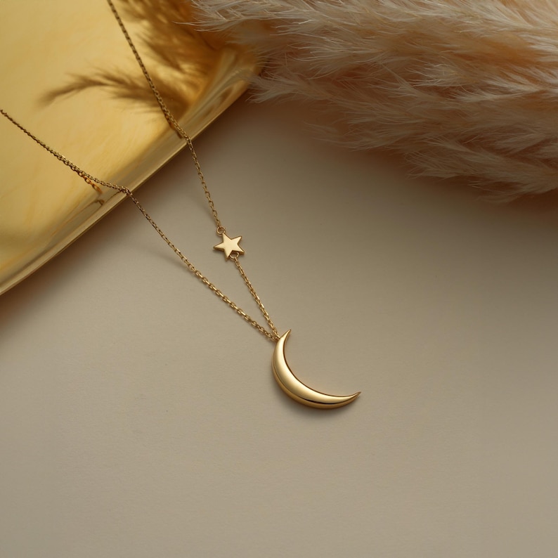 Pu&ograve; includere: Una collana d'oro con un ciondolo a forma di luna crescente e un piccolo ciondolo a forma di stella. La collana &egrave; su uno sfondo bianco.
