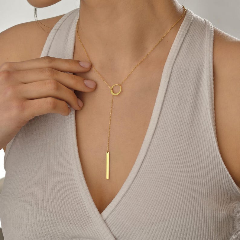May include: A gold necklace with a circle pendant and a long, thin bar pendant hanging from it.