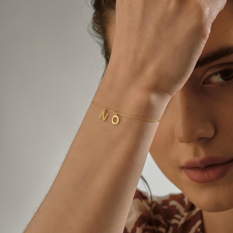 K&ouml;nnte beinhalten: Ein goldenes Kettenarmband mit den Buchstaben "NO", die daran h&auml;ngen. Das Armband befindet sich am Handgelenk einer Person.
