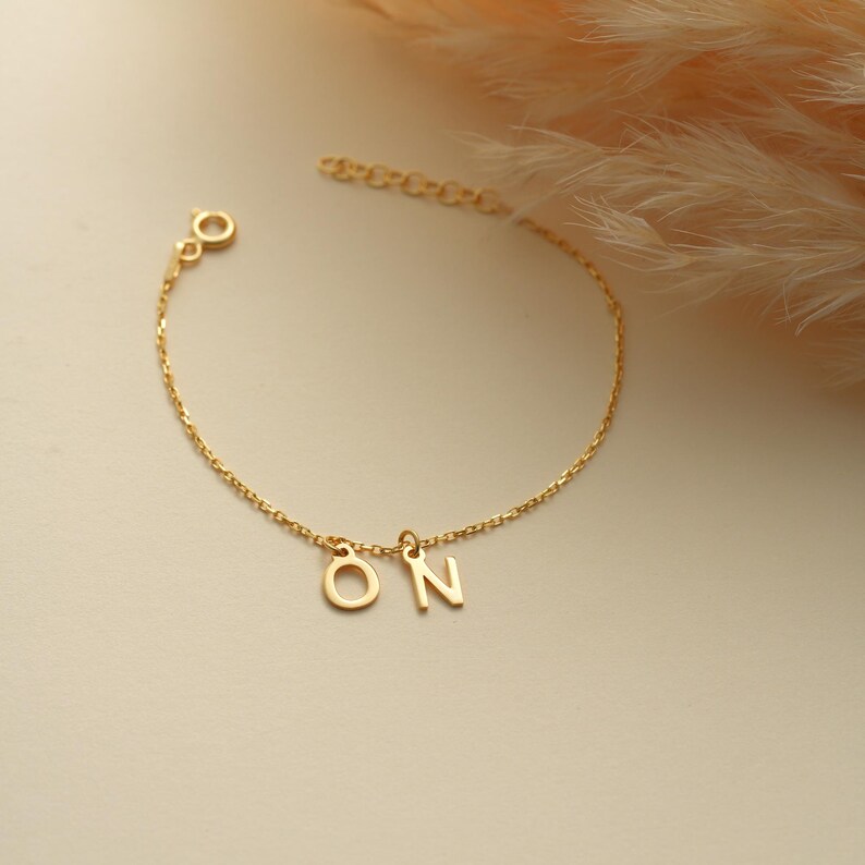 K&ouml;nnte beinhalten: Ein goldenes Kettenarmband mit den Buchstaben "ON", die daran h&auml;ngen.