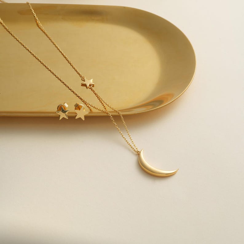 Pu&ograve; includere: Una collana d'oro con un ciondolo a forma di luna crescente e due orecchini a forma di stella. I gioielli sono esposti su un vassoio d'oro.