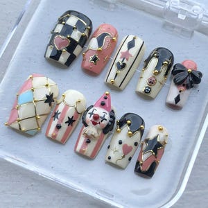May include: A set of ten press-on nails featuring a circus theme with black, white, pink, and blue colors. Designs include a checkered pattern, a 3D clown face, a black bow, stars, hearts, stripes, and gold metallic studs on a glossy finish.