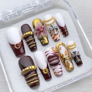 May include: A set of ten press-on nails in a clear plastic case, featuring a mix of deep burgundy and white colours. The designs include gold metallic bands, a 3D flower, silver cross charms, gold bead chains, and a 3D set of lips on a white base.