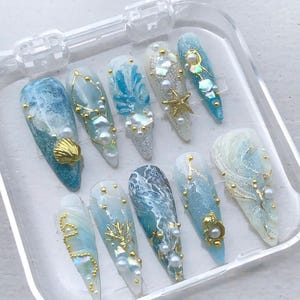 May include: A set of ten stiletto-shaped press-on nails in a clear plastic case. The nails feature ocean-inspired designs with shades of blue, white, and gold, decorated with small pearls, gold metal charms, starfish, and iridescent shell pieces.
