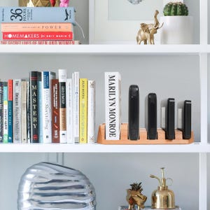 May include: A white bookshelf with books and decorative items. The top shelf has a gold elephant figurine and a potted plant. The middle shelf has a variety of books, including "Marilyn Monroe" and "The Girl on the Train". The bottom shelf has a silver bowl, a gold watering can, and a gold pineapple figurine.