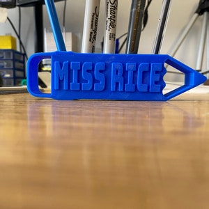 May include: A blue plastic pencil holder with the name "MISS RICE" printed on it. The holder has a slot for pens and pencils and is shaped like a pencil.