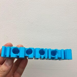 May include: A bright blue 3D printed object featuring a series of four circular openings along its length. The structure has a layered, textured surface finish and a rectangular block-like design with vertical segments between each circular hole.