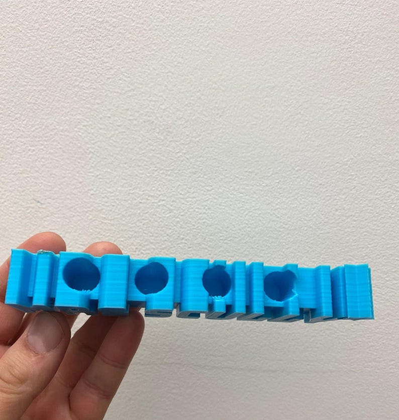 May include: A bright blue 3D printed object featuring a series of four circular openings along its length. The structure has a layered, textured surface finish and a rectangular block-like design with vertical segments between each circular hole.