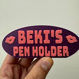 Könnte beinhalten: Lilafarbener Stifthalter mit dem Text "BEKI'S PEN HOLDER" in rosa Buchstaben und zwei rosa Lippen auf jeder Seite.