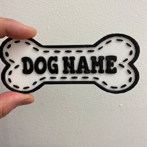以下が含まれることがあります： 黒い輪郭とステッチのデザインが施された白いプラスチック製の犬の骨のタグ。タグには黒い文字で「DOG NAME」と書かれています。